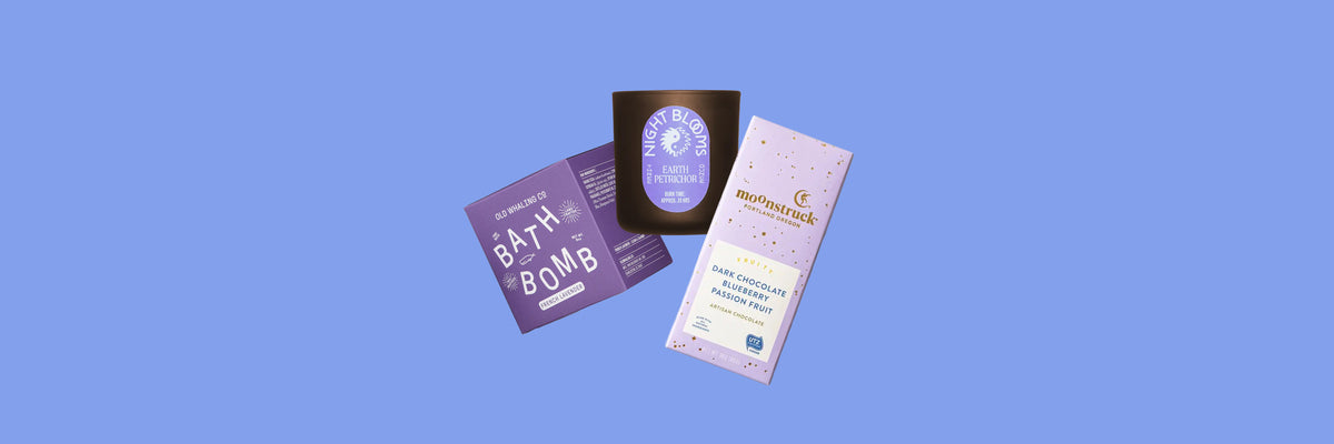 Purple bath bomb box, brown glass candle and lavender chocolate bar on a periwinkle blue background