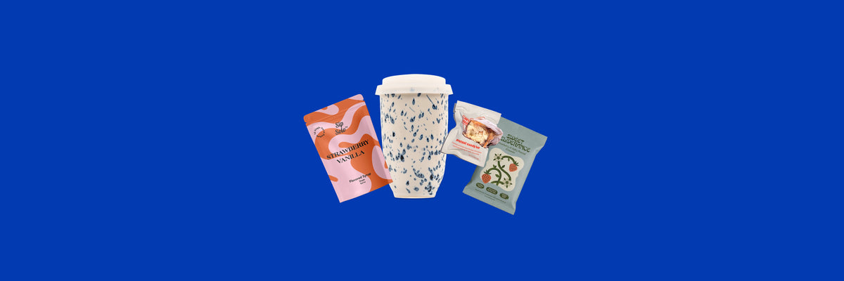 Strawberry vanilla syrup pouch, speckled travel cup, candy bar and granola bag on a cobalt blue background
