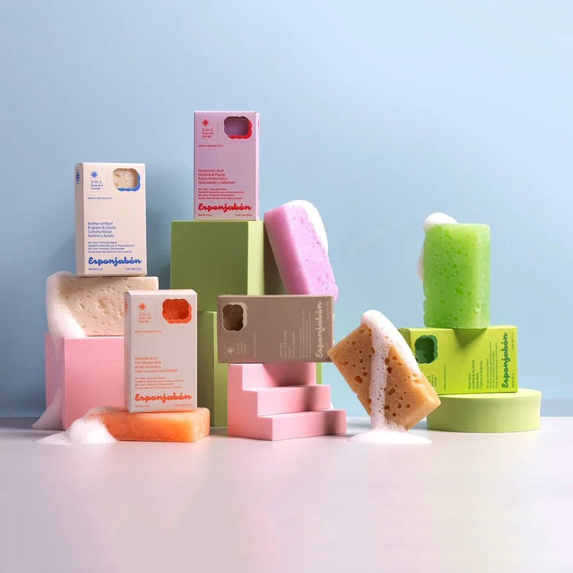 A styled product display of Esponjabon bath sponge bars and their matching packaging boxes, arranged on pastel pink and green blocks against a soft light blue background.