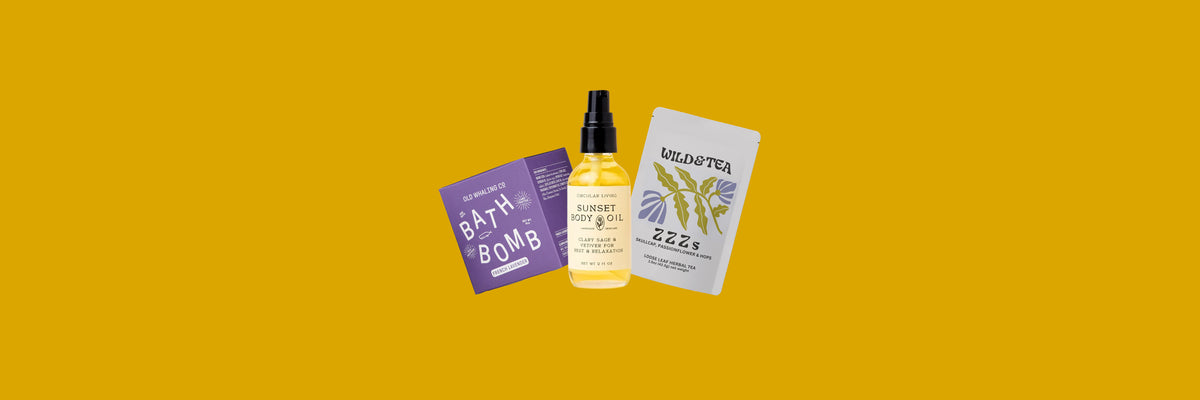 Purple bath bomb box, body oil bottle and gray tea pouch on a mustard yellow background