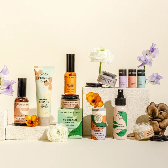 Good Flower Farm skincare on white pedestals against warm cream, styled with white and purple flowers and mushrooms.