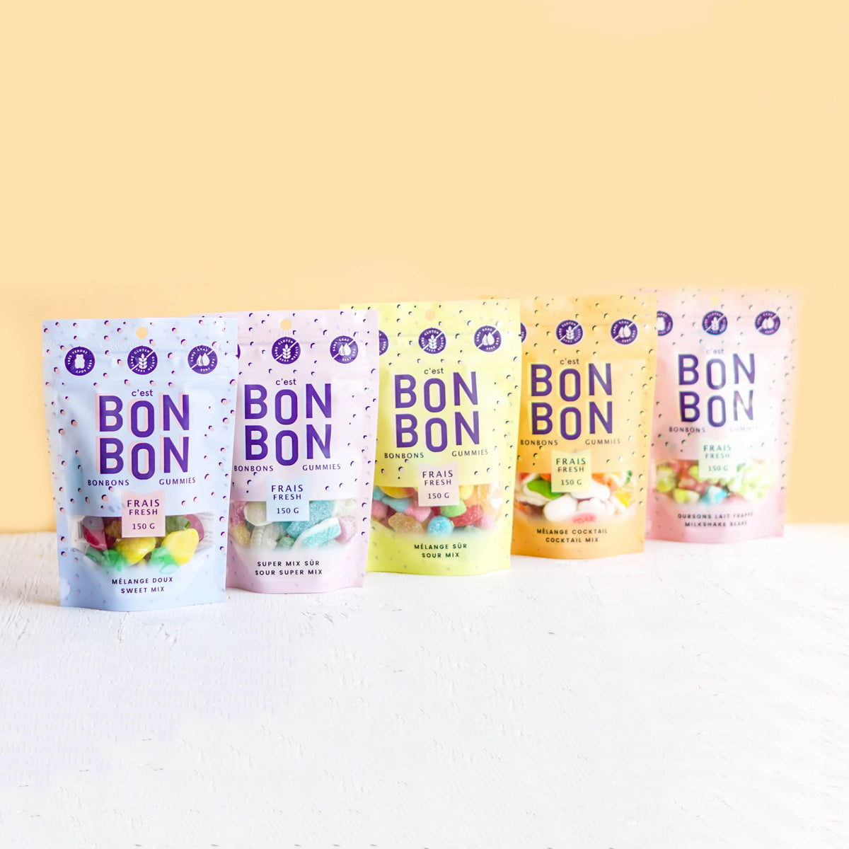 Five stand-up pouches of Bon Bon candy in assorted fruit flavors are lined up on a white textured surface against a pale yellow background. The colorful packaging features purple 'BON BON' branding and different pastel color schemes for each flavor.