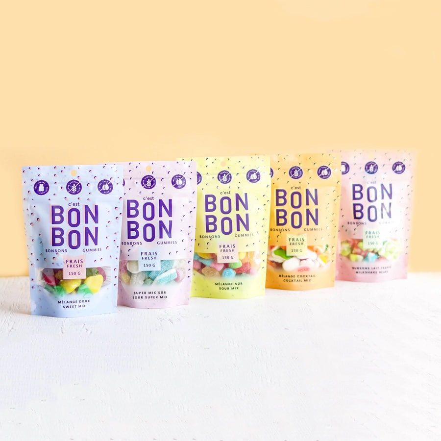 Five stand-up pouches of Bon Bon candy in assorted fruit flavors are lined up on a white textured surface against a pale yellow background. The colorful packaging features purple 'BON BON' branding and different pastel color schemes for each flavor.