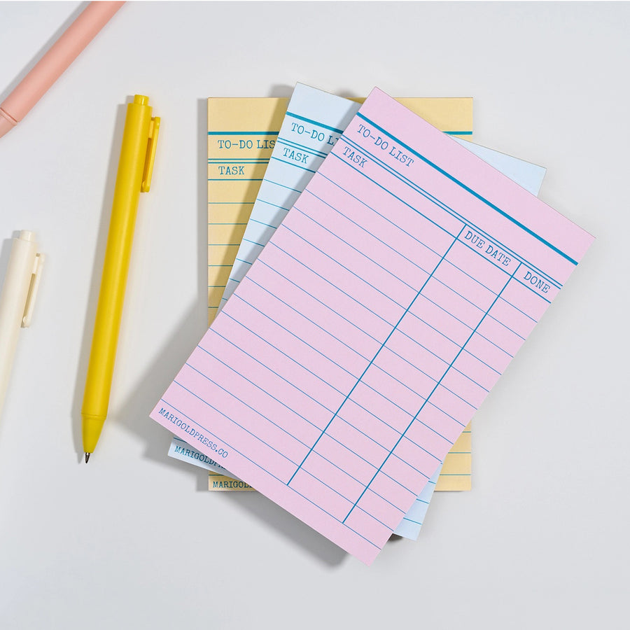 A stack of pastel notepad sheets with lined to-do lists and date columns, arranged on a light gray surface with a yellow pen and partial pastel pens nearby.