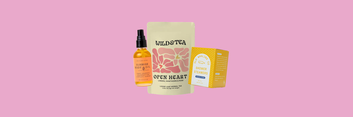 Orange body oil bottle, rose-colored tea pouch and yellow shower steamers box on a pink background