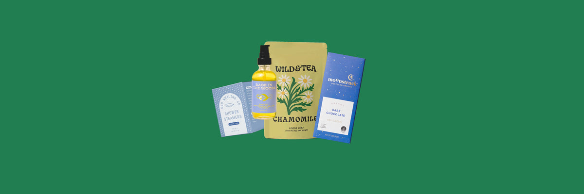 Blue shower steamers box, body oil bottle, chamomile tea pouch and a blue chocolate bar on a green background