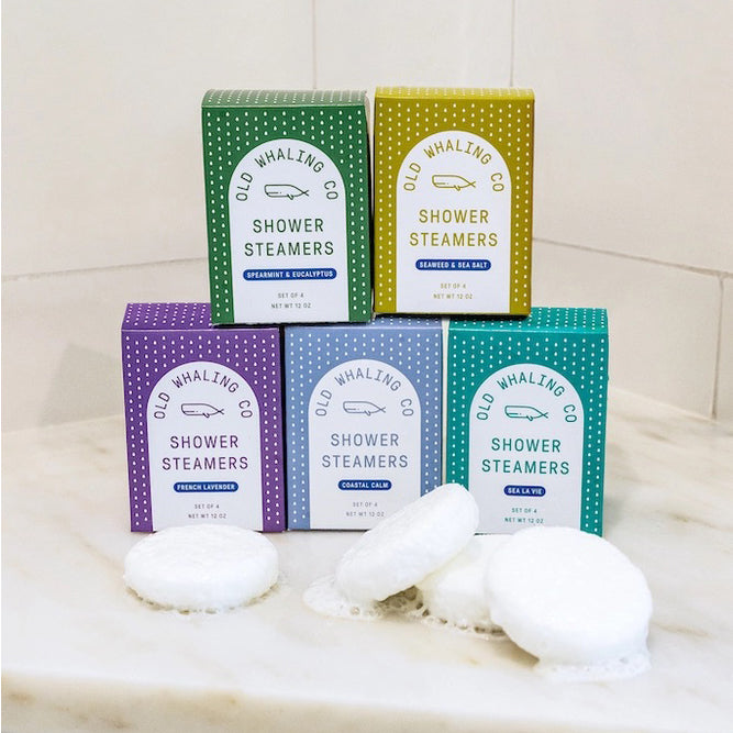 Five boxes of Old Whaling Co. Shower Steamers in green, yellow, purple, light blue and teal on marble, with white steamer pads in front.