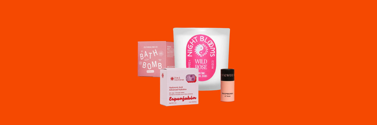 Pink bath bomb box, white candle, soap and sponge box and lip balm tube on an orange background