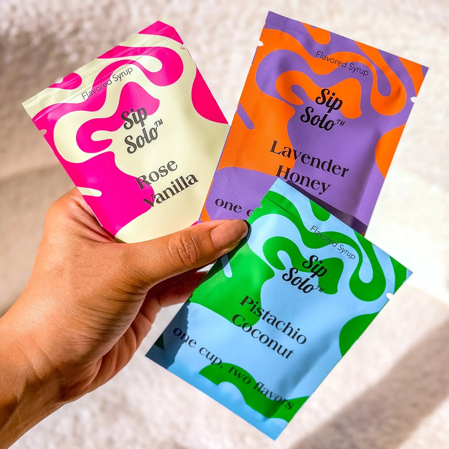 A hand holds three Sip Solo flavored syrup sachets against a soft light background. The sachets are pink and cream (Rose Vanilla), orange and purple (Lavender Honey), and green and light blue (Pistachio Coconut).